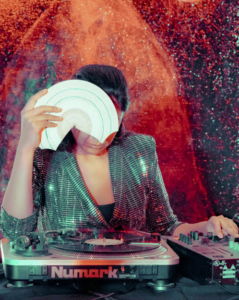 Maria Chavez stands at a turntable and holds a broken plate with her right hand in front of her face. She wears a sparkling green blazer.